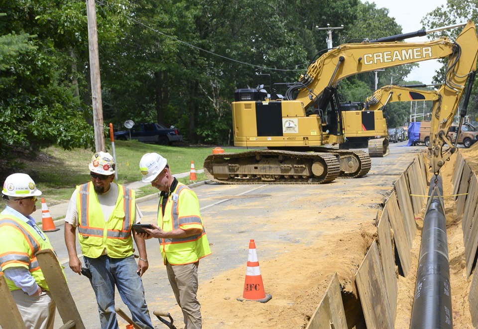 Three coworkers talk on a job site for a gas pipeline along a road.